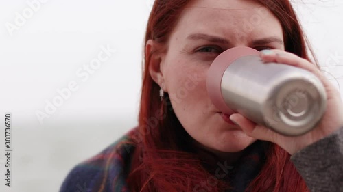 Beautiful young girl with long red hair drinking hot coffee or teadrink from a thermo mug sitting on the sea photo