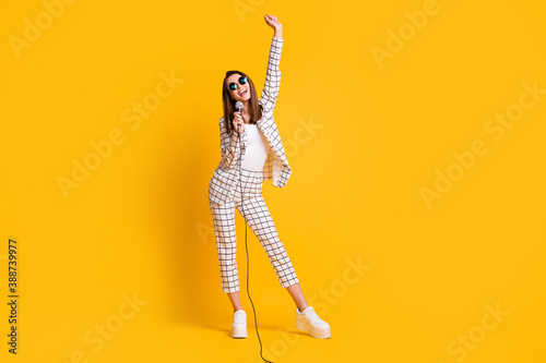 Full length body size photo of girl holding mic singing keeping hand up smiling wearing sunglass isolated on vibrant yellow color background
