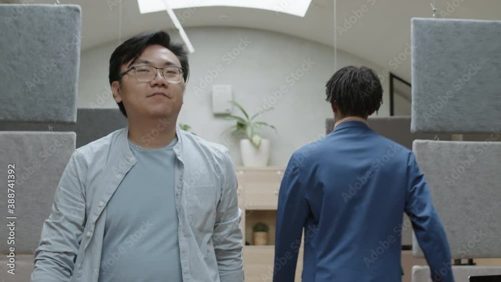 Medium shot of young Asian businessman wearing glasses going along modern office, greeting his male African colleague and then sitting down at workplace and opening laptop Medium shot of young Asian businessman wearing glasses going along modern office, greeting his male African colleague and then sitting down at workplace and opening laptop