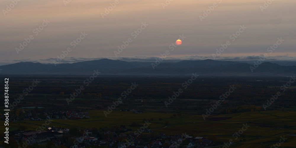 Beautiful sunset with sun shining through a veil of clouds above Rhine valley and Vosges mountains viewed from mountain range Kaiserstuhl with small village Vogtsberg in autumn.