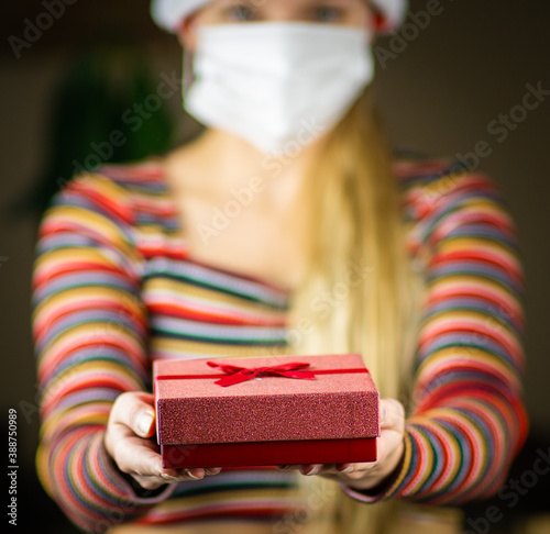 Obraz na płótnie Young blonde caucasian girl in Santa hat and striped jumper , with protective mask on face, holding, giving a gift, present to the camera. Coronavirus lockdown Christmas concept. w salonie