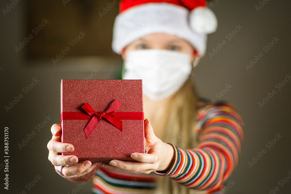 Young blonde caucasian girl in Santa hat and striped jumper , with protective mask on face, holding, giving a gift, present to the camera. Coronavirus lockdown Christmas concept.