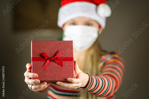 Obraz na płótnie Young blonde caucasian girl in Santa hat and striped jumper , with protective mask on face, holding, giving a gift, present to the camera. Coronavirus lockdown Christmas concept. w salonie