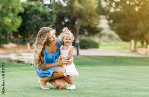 happy Mom and her child baby daughter sitting in the grass and having fun togetherss in sunny day outdoors, wearing dresses, sunset light, copy space