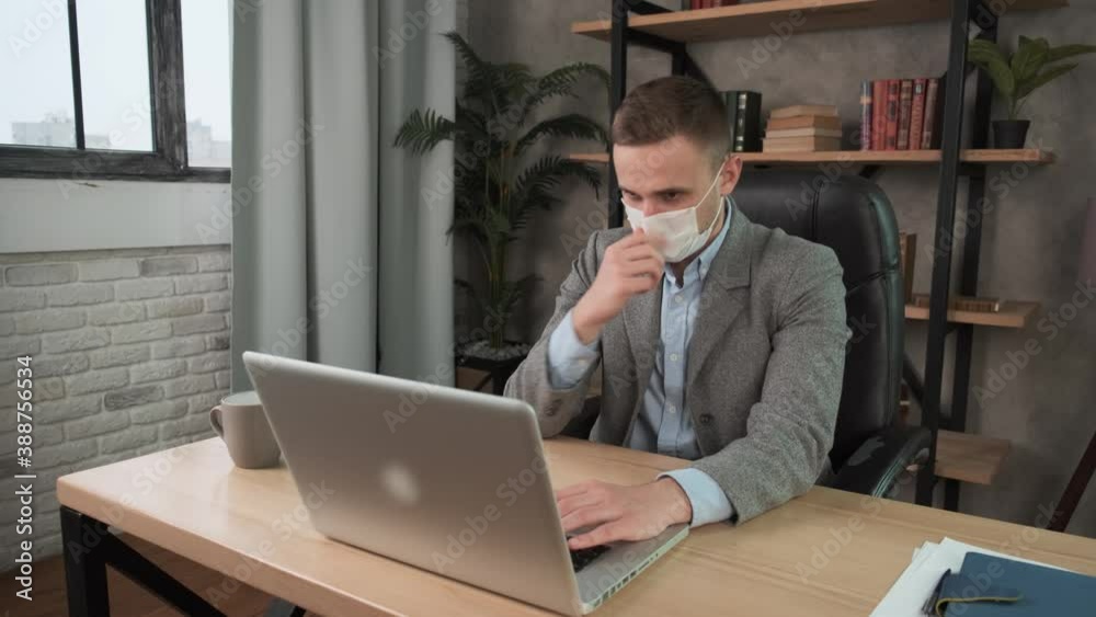 Young confident man employee in a medical facial mask works in the office at the computer. Male worker looking at camera and showing thumb down, working remotely at home. Work during self-isolation Young confident man employee in a medical facial mask works in the office at the computer. Male worker looking at camera and showing thumb down, working remotely at home. Work during self-isolation
