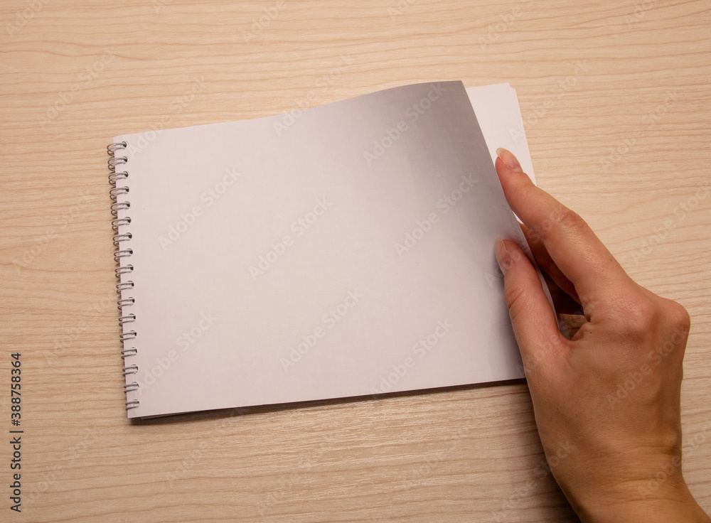 Female left hand lies on a white blank notebook against a wooden background Female left hand lies on a white blank notebook against a wooden background