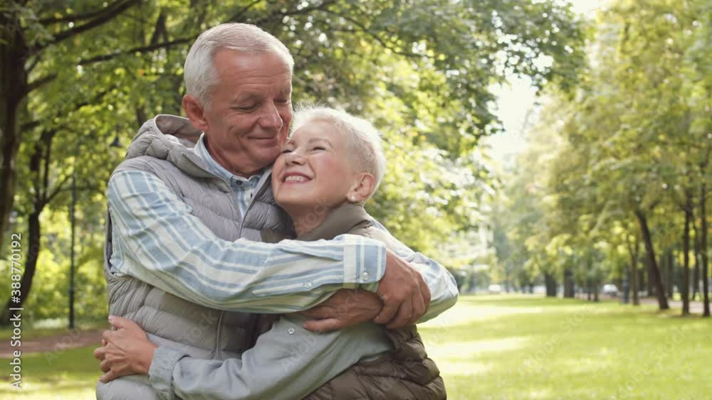 Medium arc POV of loving senior Caucasian man and short-haired elderly woman standing in green public park looking in sky then into eyes of each other, hugging and smiling on camera Medium arc POV of loving senior Caucasian man and short-haired elderly woman standing in green public park looking in sky then into eyes of each other, hugging and smiling on camera
