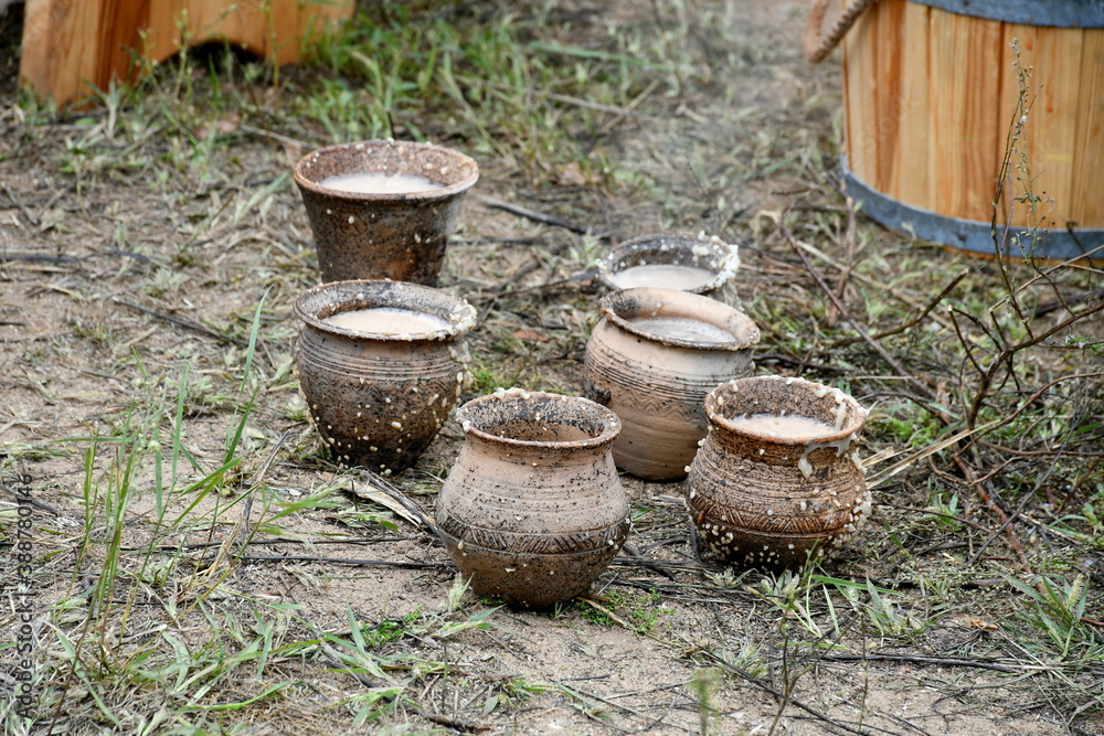 A close up on a set of freshly made clay pots, cups, and mugs with some milk and yeast in them to help them solidify seen next to a bucket of water on a grassy soil spotted on a cloudy summer day A close up on a set of freshly made clay pots, cups, and mugs with some milk and yeast in them to help them solidify seen next to a bucket of water on a grassy soil spotted on a cloudy summer day