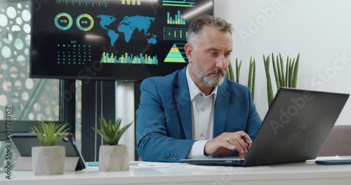 Handsome concentrated confident 50-aged bearded businessman in formal clothes sitting in front of computer and working over business project in his office photo