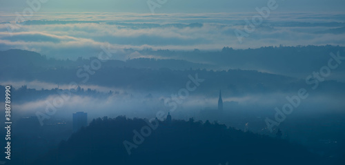 Amazing foggy sunrise over the city of Graz with Schlossberg hill and Church of the Sacred Heart of Jesus tower, in Styria region, Austria. Panoramic view from Plabutsch mountain on autumn morning.
