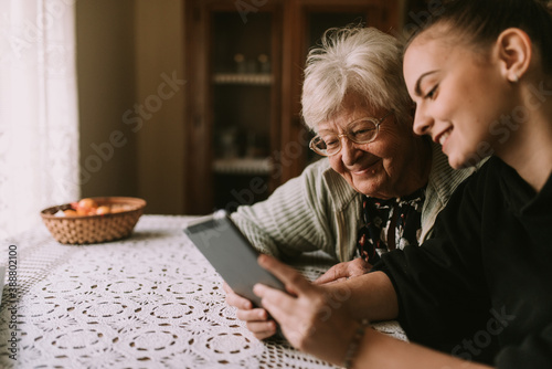 A cute gray-haired retired grandmother and her teenage caucasian granddaughter look at a tablet as they sit at a table in their grandmother's house. Grandma's education about new technology