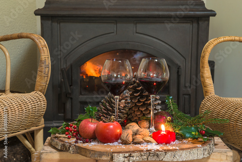 Winter holidays still life. Two glasses of red wine, fruits, nuts, cones, fir branches on the plate in front of the burning stove © stsvirkun