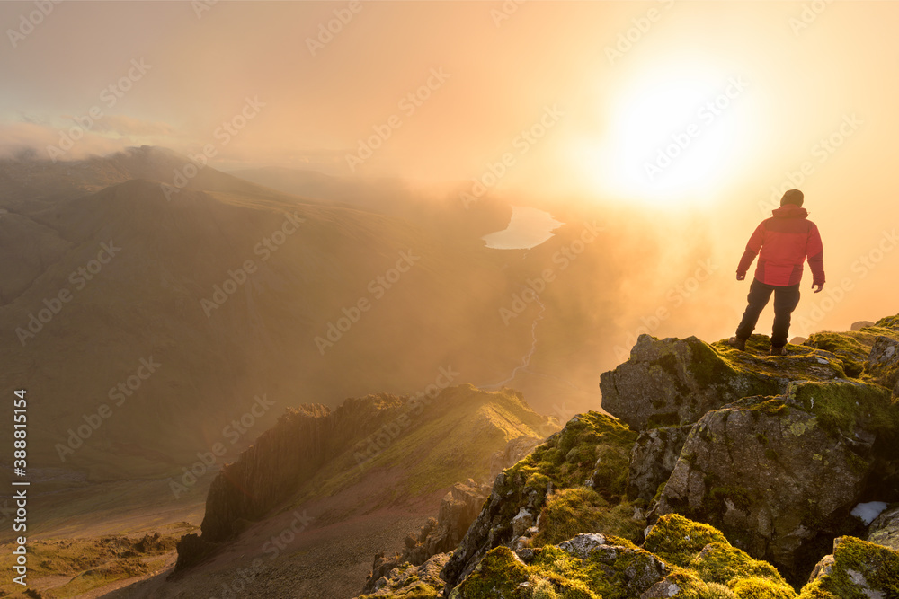 Fototapeta Lone hiker wearing red coat on mountain top with misty dramatic view. Taken in the Lake District, UK.