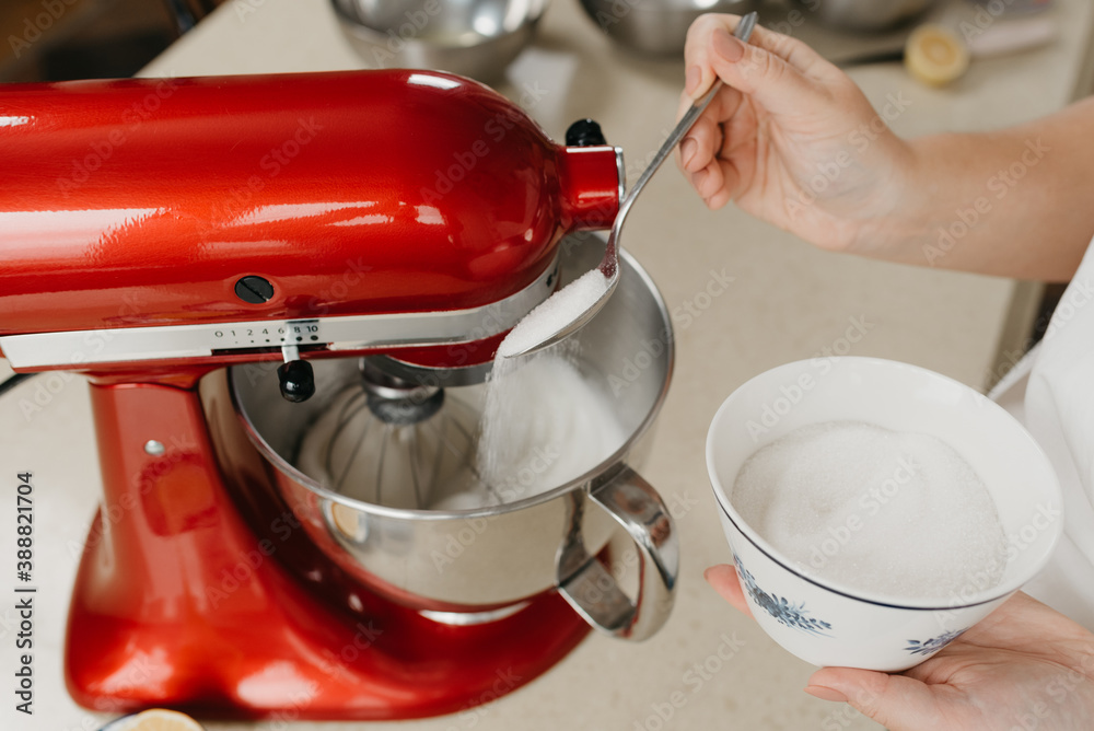 A close photo of the hands of a young woman who is spooning sugar to stainless steel soup bowl with the egg whites while the red stand mixer is whipping it.
