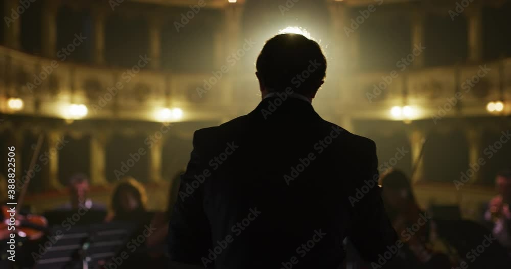 Cinematic close up shot of conductor directing symphony orchestra with performers playing violins, cello and trumpet on classic theatre with curtain stage during music concert with dramatic lights.