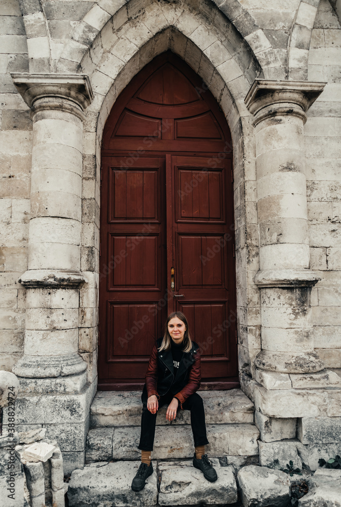 young woman in a leather jacket near the entrance to the ancient wooden door of a gothic temple