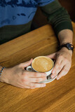 Male's hands holding a cup of cappuccino coffee, wooden table