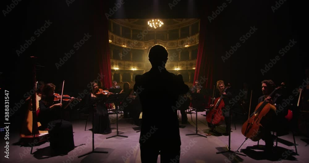 Cinematic shot of conductor directing symphony orchestra with performers playing violins, cello and trumpet on classic theatre with curtain stage during music concert with dramatic lights.