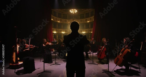 Cinematic shot of conductor directing symphony orchestra with performers playing violins, cello and trumpet on classic theatre with curtain stage during music concert with dramatic lights. photo
