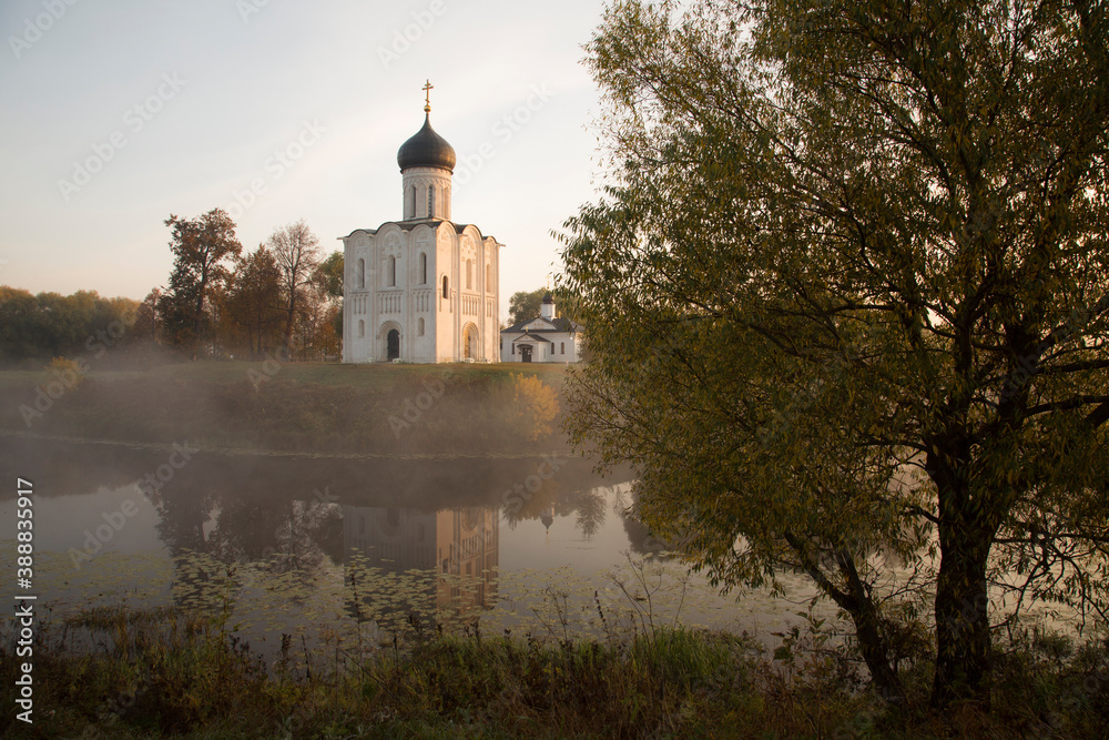 An XII century Russian Orthodox church above a misty pond lit by the rising sun. A tree in the foreground. Autumn colours. No people. An XII century Russian Orthodox church above a misty pond lit by the rising sun. A tree in the foreground. Autumn colours. No people.