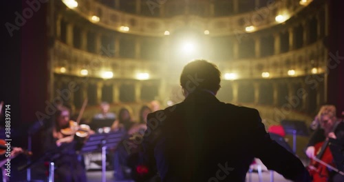 Cinematic shot of conductor directing symphony orchestra with performers playing violins, cello and trumpet on classic theatre with curtain stage during music concert with dramatic lights.