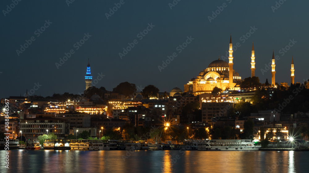 Naklejka premium Istanbul-September 23, 2020. Evening view of the Suleymaniye mosque, across the Golden horn Bay. Mosque and buildings on the shore in the lights that are reflected in the night water of the Bay.
