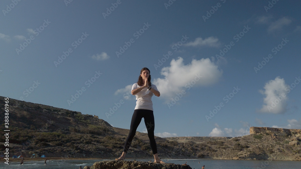 Obraz premium Young caucasian woman brunette doing yoga on rocky beach against blue sky. High quality photo