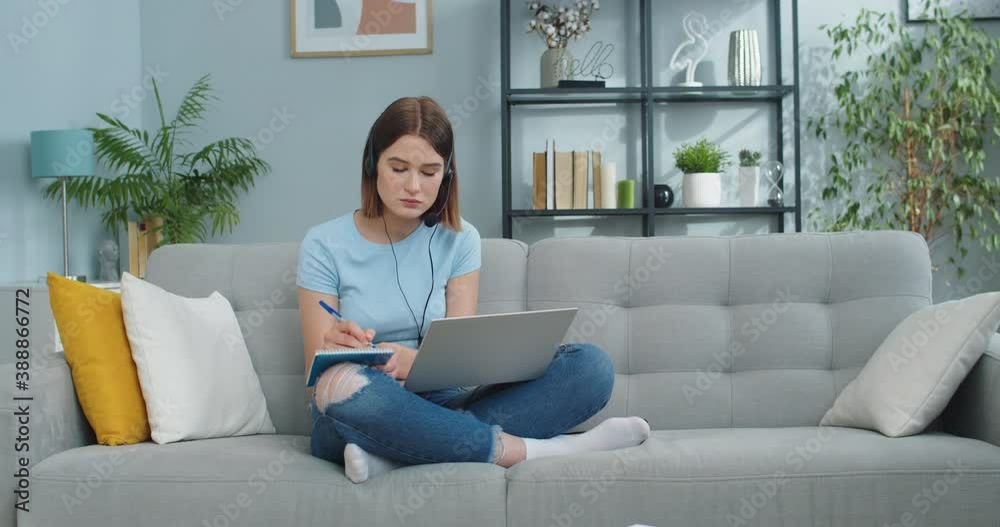 Smart Caucasian female student in headphones studying at home during pandemic period. Good-looking young woman working on laptop and taking notes while listening to lecture. Online education. Smart Caucasian female student in headphones studying at home during pandemic period. Good-looking young woman working on laptop and taking notes while listening to lecture. Online education.