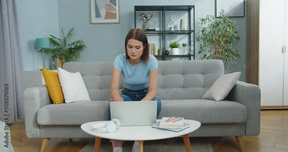 Pretty Caucasian female student sitting on sofa and studying on laptop at home. Attractive young woman working and typing in living room indoors. Technology, Internet, education concept. Pretty Caucasian female student sitting on sofa and studying on laptop at home. Attractive young woman working and typing in living room indoors. Technology, Internet, education concept.