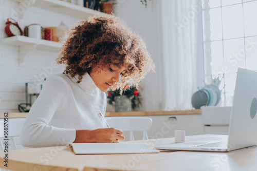 Fotografie An African-American young girl with a buxom hairstyle takes online training at home sitting in the kitchen in front of a laptop and records a lecture on a Sunny day