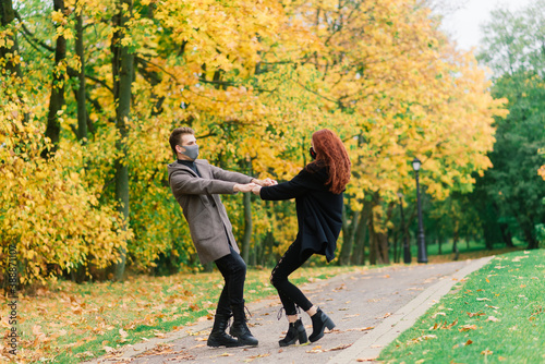 Young red haired woman puts on face mask while walking with young man in autumn park.