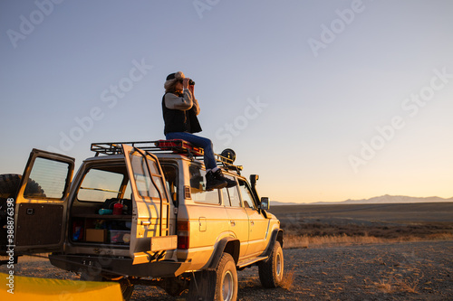 young girl traveler sits on the roof of a large jeep car and looks through binoculars photo