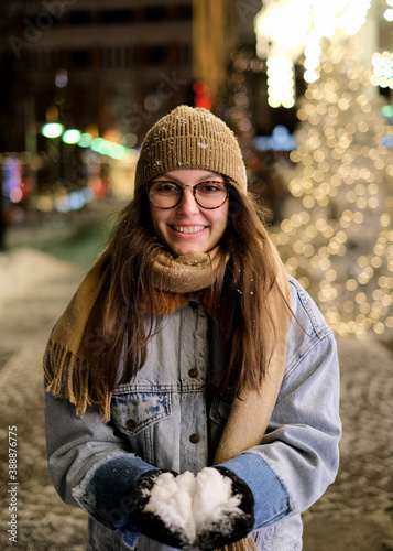 Cheerful woman with a scarf and gloves, blowing snowflakes from her hands, outdoors, on the street, surrounded by coloured bokeh. Winter and christmas concept. Shallow deep of field. photo