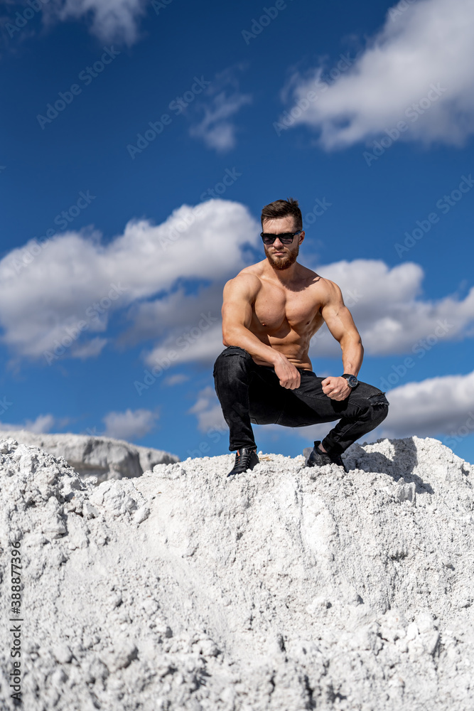 Man with perfect body pumping up muscles. Sitting in a quarry. Grey rocks background. Man with perfect body pumping up muscles. Sitting in a quarry. Grey rocks background.