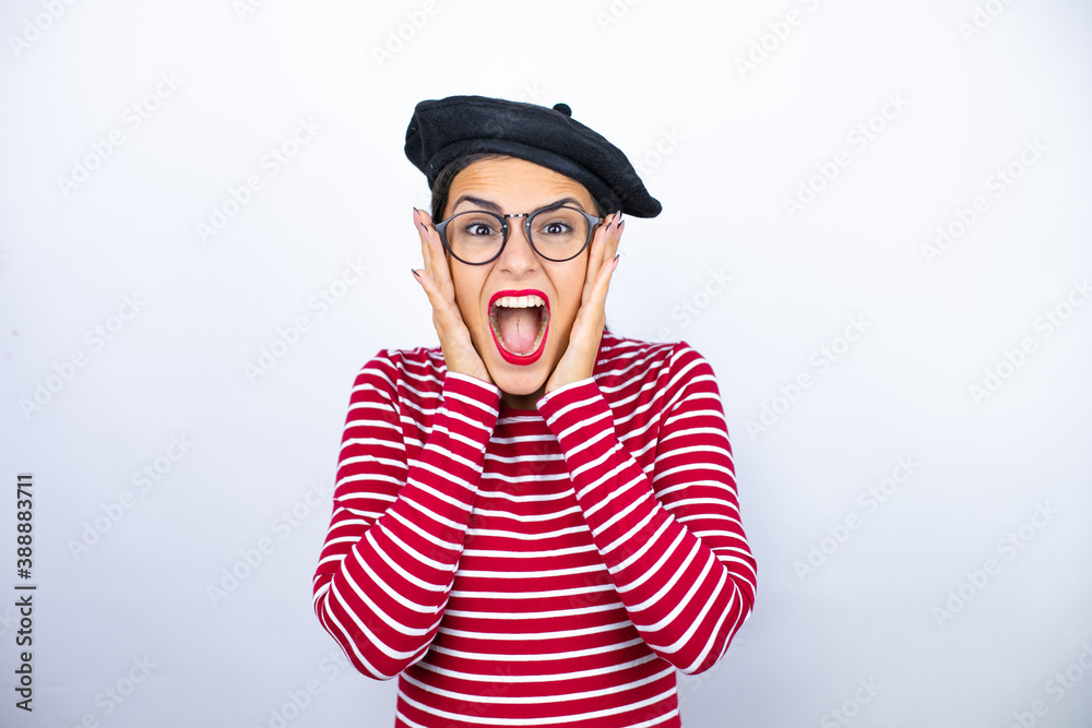 Fototapeta premium Young beautiful brunette woman wearing french beret and glasses over white background crazy and scared with hands on head, afraid and surprised of shock