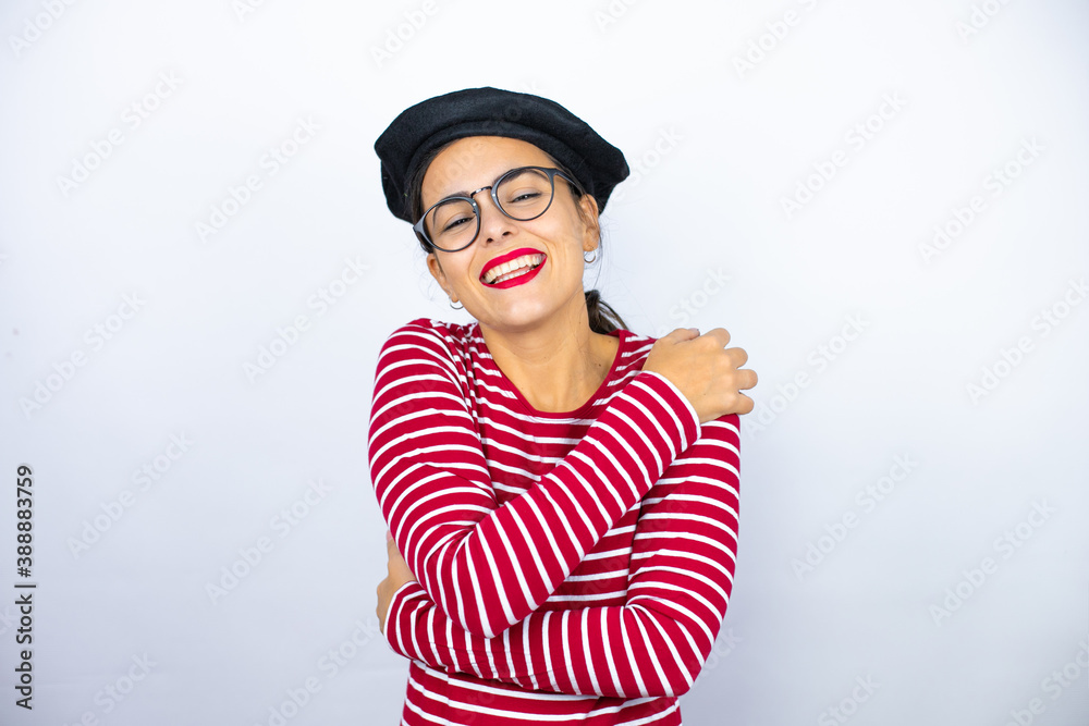 Young beautiful brunette woman wearing french beret and glasses over white background hugging oneself happy and positive, smiling confident Young beautiful brunette woman wearing french beret and glasses over white background hugging oneself happy and positive, smiling confident