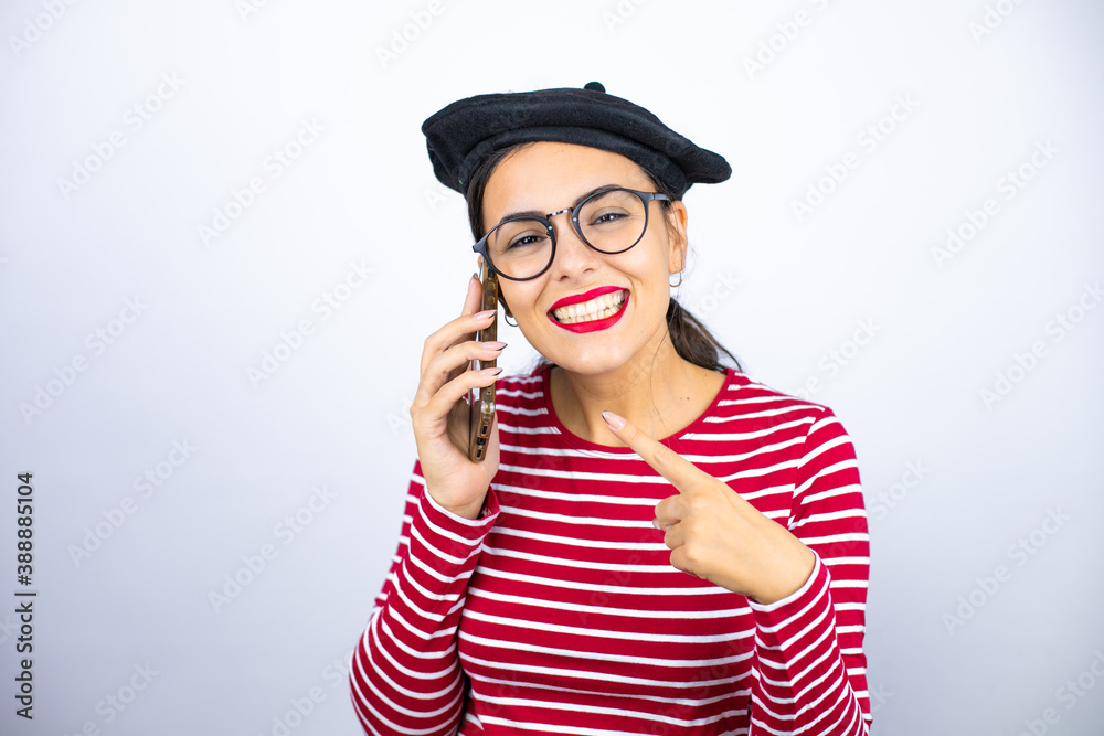 Young beautiful brunette woman wearing french beret and glasses over white background smiling, pointing to the phone that he has in his ear Young beautiful brunette woman wearing french beret and glasses over white background smiling, pointing to the phone that he has in his ear
