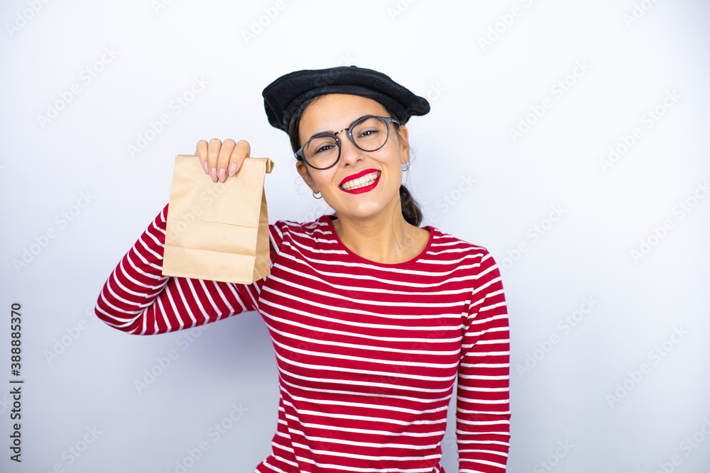 Young beautiful brunette woman wearing french beret and glasses over white background smiling and holding a paper bag