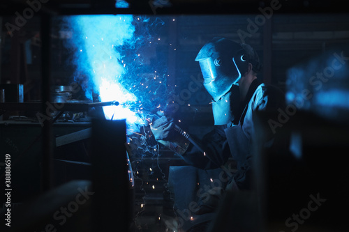 Industrial worker in protective mask using modern welding machine for welding metal construction on production manufacture at metalworking factory