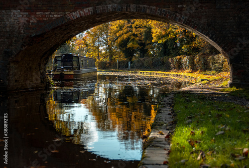 a view of a narrow boat under the arch of a brick foot bridge reflected into the still water of the canal, with autumn colours in the trees photo