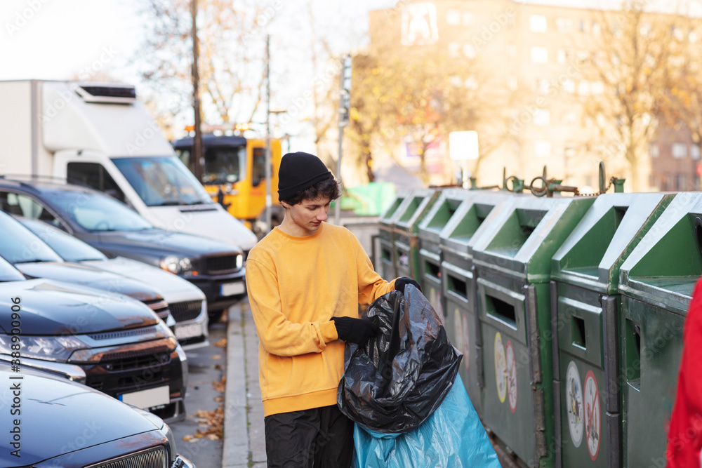 Young male volunteer holding waste in plastic bag by garbage can Young male volunteer holding waste in plastic bag by garbage can