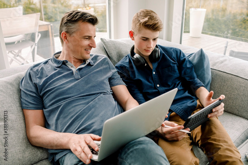 Smiling father with laptop looking at son using digital tablet while sitting on sofa at home photo