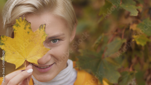 Portrait shot of young caucasian blonde woman with blue eyes hiding face with yellow autumn leaf. High quality photo