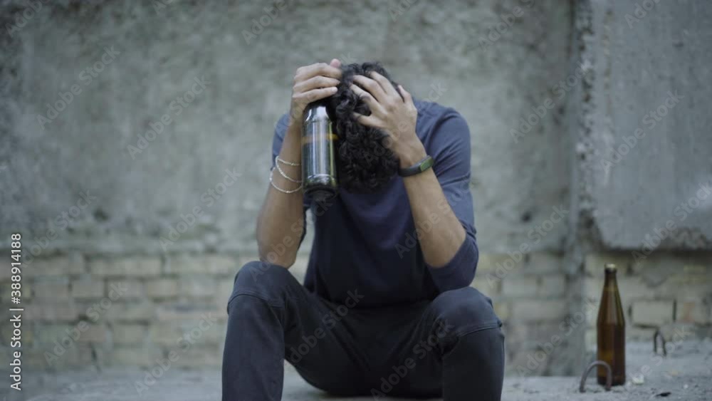 Desperate crying young man drinking beer on urban ruins. Portrait of frustrated mixed-race addicted guy in ghetto. Social inequality and alcohol addiction concept.