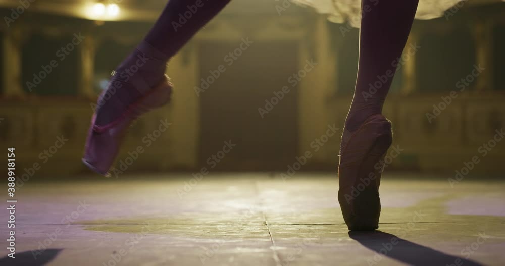 Cinematic close up shot of an young graceful classical ballet female dancer feet in pointe shoes and white tutu is performing a choreography on classic theatre stage with dramatic lighting before sta