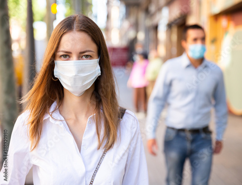 Focused young woman in medical mask walking to work along city street on spring day. New life reality during coronavirus pandemic.