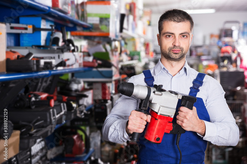 Confident man in blue coverall is choosing drilling machine in tools shop