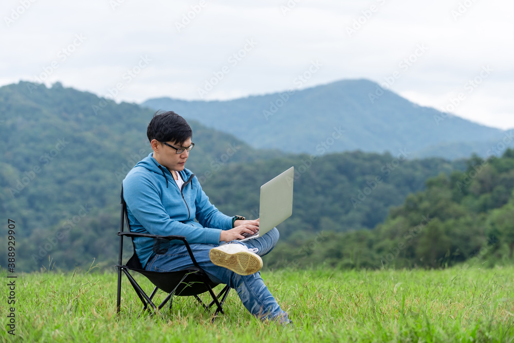 Asian man sitting on the chair using laptop computer with internet for online working or video conference on the mountain. Handsome guy relax and enjoy outdoor lifestyle in autumn holiday vacation.