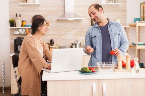 Couple looks at recipe for organic salad on laptop in kitchen. Man helping woman to prepare healthy organic dinner, cooking together. Romantic cheerful love relationship