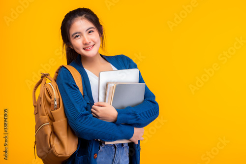 Happy asian female college student smiling at camera on yellow background and copy space, holding laptop and books, hanging bagpack. Youth girl student is exchange student. education concept